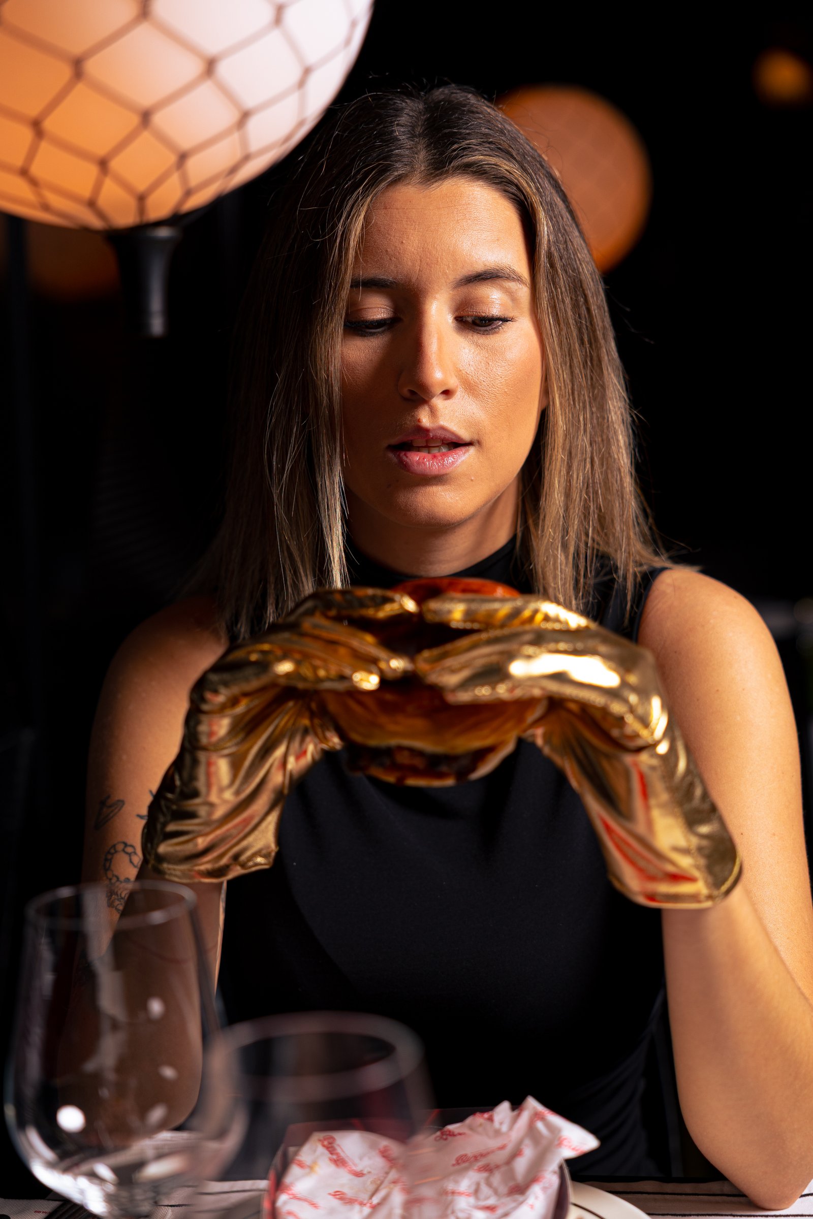 Mujer con guantes dorados sosteniendo una hamburguesa Burgaccia con luces cálidas de fondo — fotografía lifestyle de marca