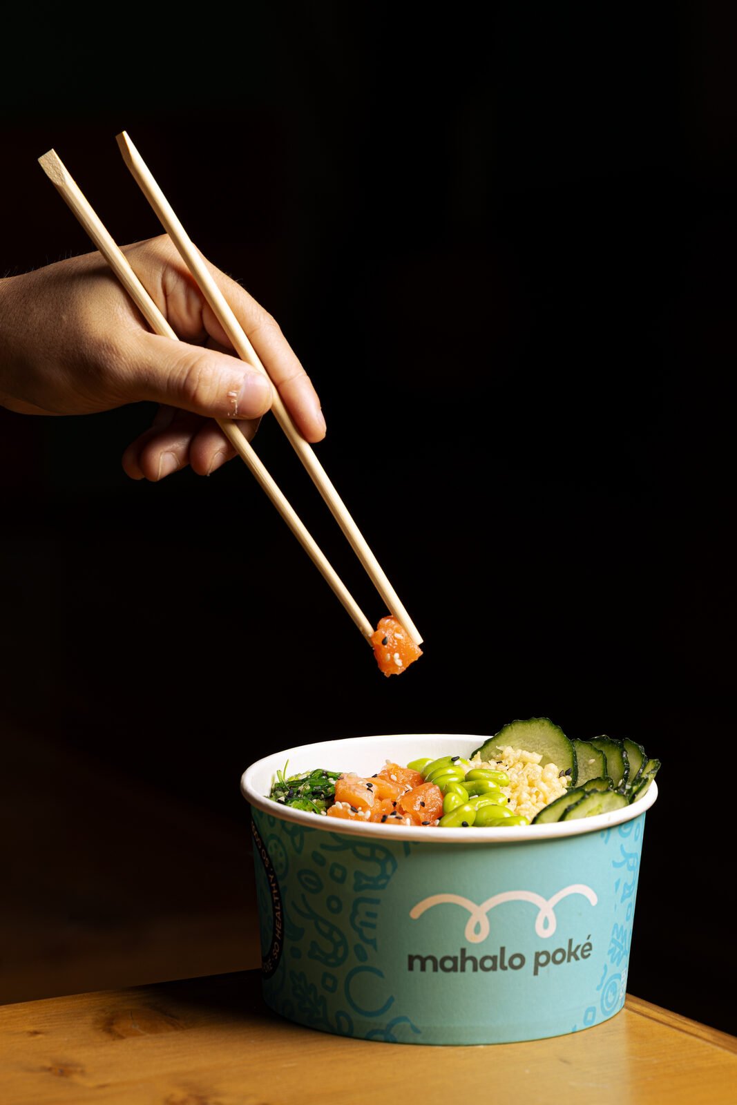 Mano con palillos levantando un cubo de salmón sobre bowl Mahalo en sesión editorial